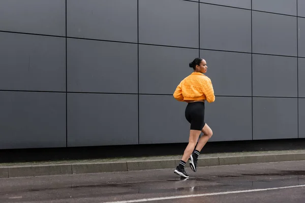 Full length of brunette african american sportswoman in bike shorts and yellow puffer jacket jogging outside - foto de stock
