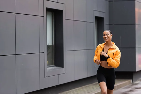 Positive african american sportswoman in bike shorts and yellow puffer jacket jogging  outside - foto de stock