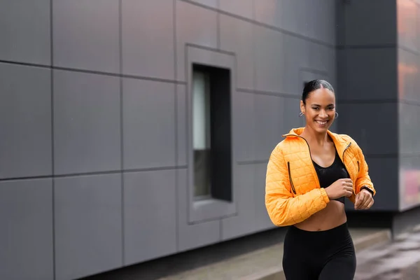 Positive african american sportswoman in yellow puffer jacket jogging outside - foto de stock