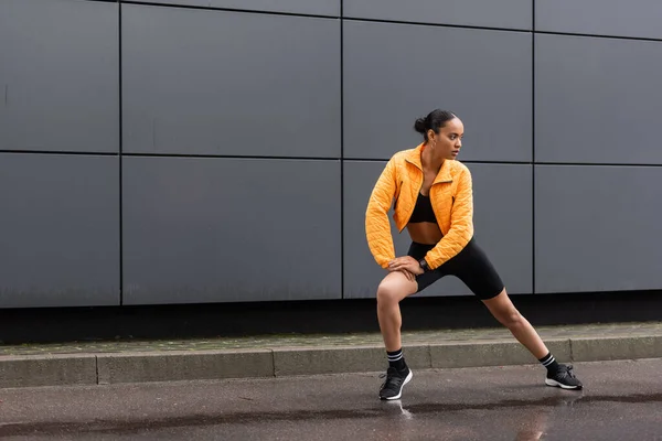 Full length of brunette african american sportswoman in bike shorts and yellow puffer jacket exercising outside — Stock Photo