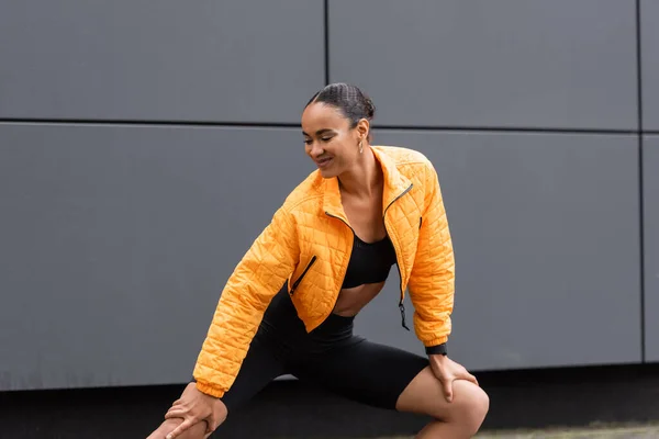 Cheerful african american sportswoman in bike shorts and yellow puffer jacket exercising outside - foto de stock