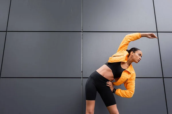 Brunette african american sportswoman in bike shorts and yellow puffer jacket exercising with hand on hip — Stockfoto