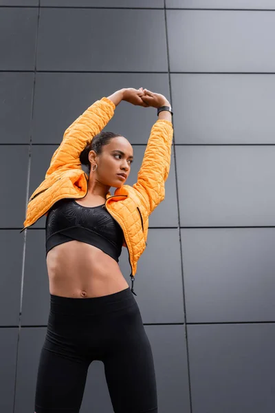 Low angle view of brunette african american sportswoman in sports bra and yellow puffer jacket warming up outside - foto de stock
