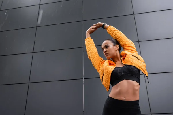 Low angle view of african american sportswoman in sports bra and yellow puffer jacket warming up outside — Fotografia de Stock