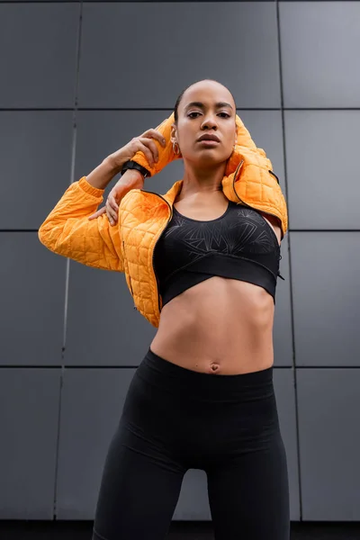 Low angle view of african american sportswoman in sports bra and yellow puffer jacket warming up near grey building — Stockfoto