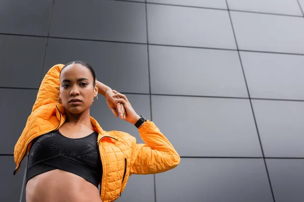 Low angle view of african american sportswoman in sports bra and puffer jacket warming up near grey building — Stock Photo