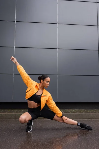 Full length of stylish african american sportswoman in bike shorts and yellow puffer jacket stretching leg near grey building - foto de stock
