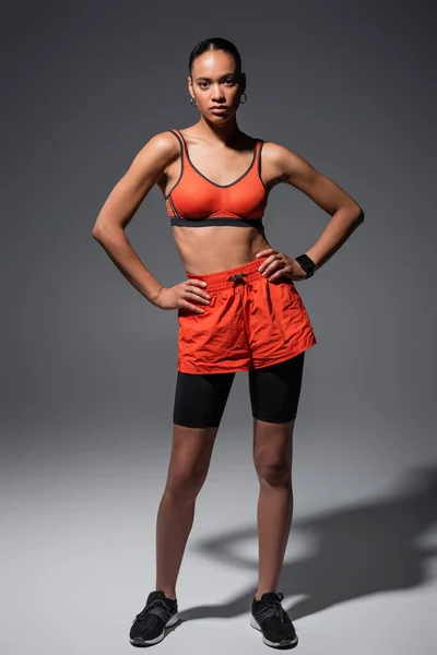 Full length of stylish african american sportswoman in bike shorts and red sports bra posing with hands on hips on grey — Stockfoto