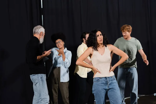 Serious multiracial woman rehearsing with hands on hips while grey haired art director talking to young actors - foto de stock