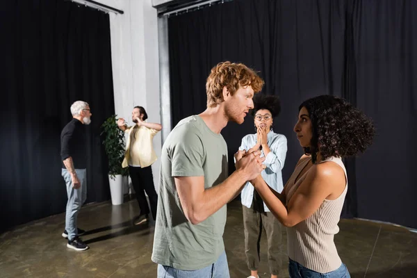 Redhead man holding hands with multiracial woman during rehearsal near interracial actors — Stockfoto