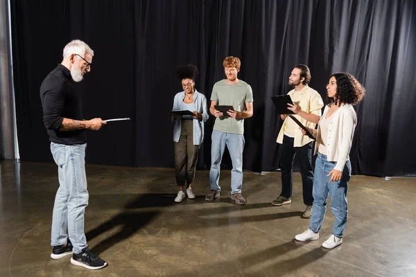Middle aged art director reading screenplay near multicultural actors in acting skills school — Stock Photo