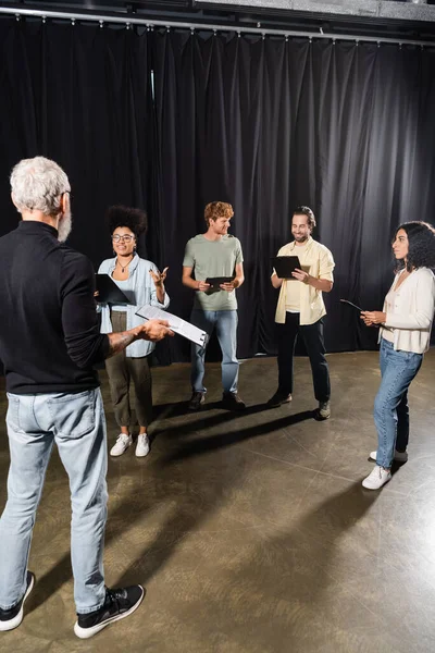 Multiethnic actors standing with screenplays near grey haired art director in theater - foto de stock