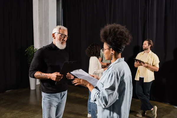 African american woman holding screenplay near bearded art director and actors in theater - foto de stock
