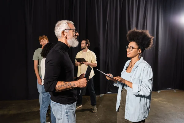 Tattooed grey haired screenwriter holding scenario near african american woman with clipboard and actors in theater - foto de stock