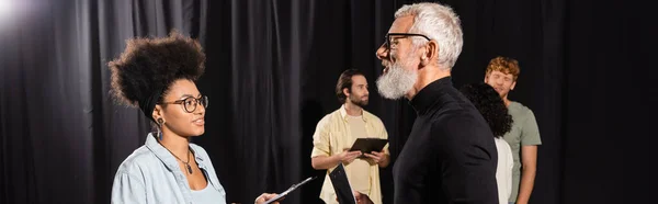 Bearded art director talking to african american actress near students on background, banner — Stockfoto