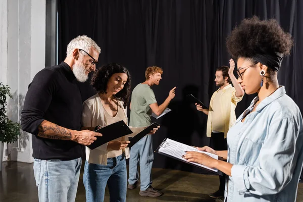 African american woman reading screenplay near bearded art director and troupe rehearsing in theater - foto de stock