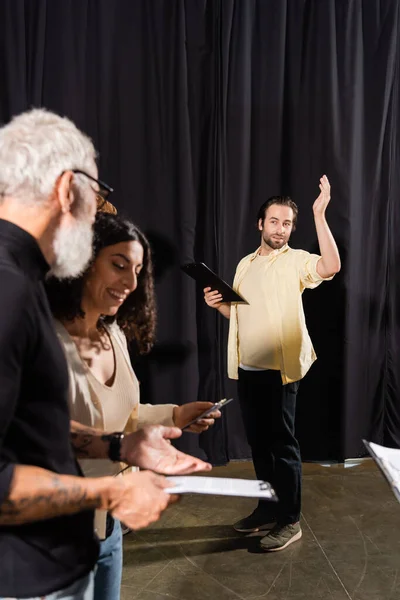 Young man with screenplay gesturing during rehearsal near smiling multiracial actress and art director on blurred foreground - foto de stock