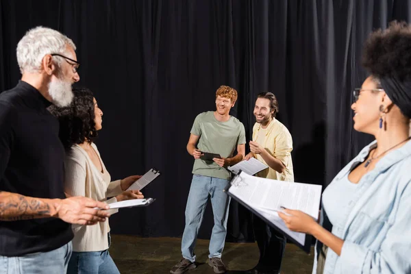 Cheerful man holding screenplay and pointing with finger near producer and actors on stage in theater - foto de stock