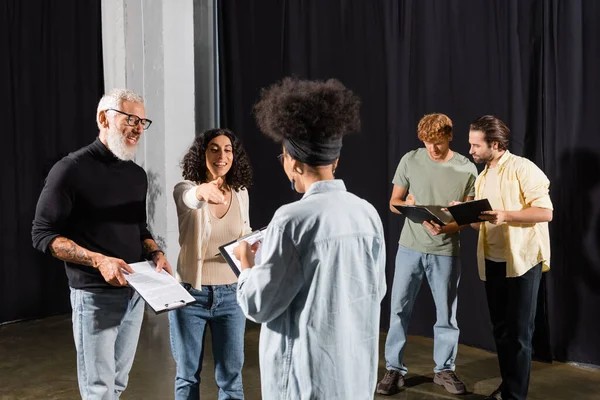 Smiling multiracial woman pointing with hand near bearded art director and african american actress on theater stage - foto de stock