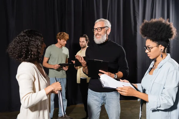 Bearded screenwriter talking to interracial actresses holding screenplays in theater school. Translation of tattoo: om, shanti, peace — Stock Photo