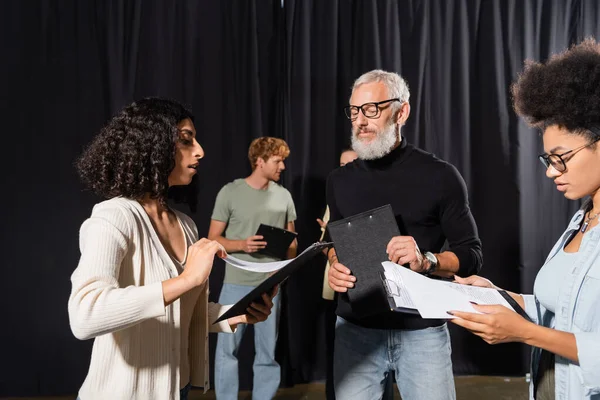 Bearded acting skills teacher looking at interracial students holding clipboards with screenplays - foto de stock