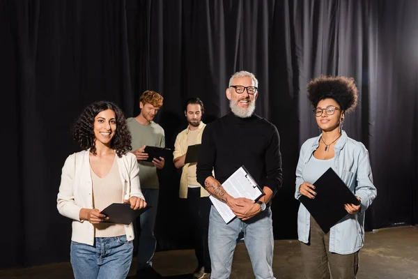 Bearded and tattooed art director with interracial actresses smiling at camera in acting studio — Stock Photo