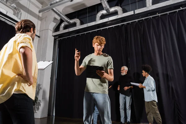 Redhead man gesturing while looking at screenplay and rehearsing near multiethnic actors in theater — Stock Photo