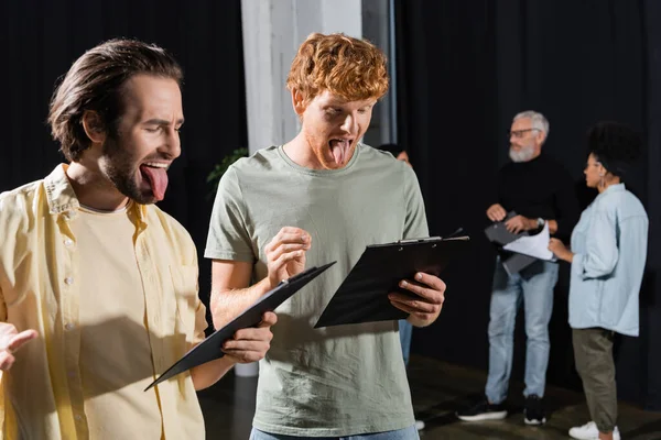 Brunette and redhead men sticking out tongues while reading scenarios during rehearsal in theater - foto de stock
