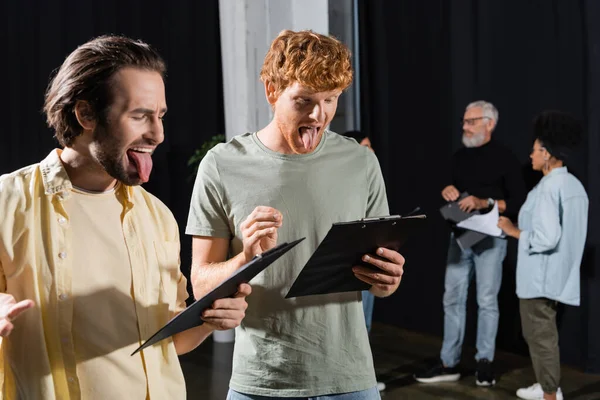Brunette and redhead men sticking out tongues while reading scenarios during rehearsal in theater — стоковое фото