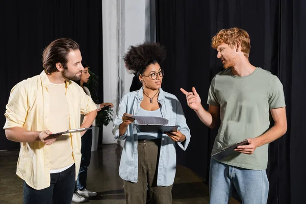 Redhead man pointing with finger while talking to interracial students with screenplays in theater - foto de stock