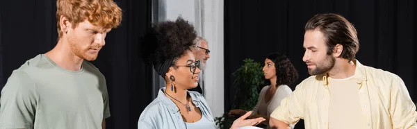 Happy african american woman talking to bearded actor near interracial students in theater studio, banner - foto de stock