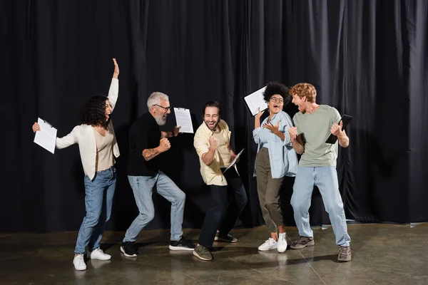 Overjoyed multiracial theater troupe holding clipboards with screenplays and showing success gesture. Translation of tattoo: om, shanti, peace — Stockfoto