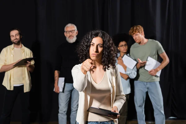 Multiracial woman with serious face expression pointing at camera while rehearsing near blurred actors and mature art director — Stock Photo