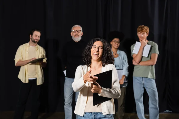 Excited multiracial actress laughing and pointing with finger near multiethnic students teacher on blurred background — стоковое фото