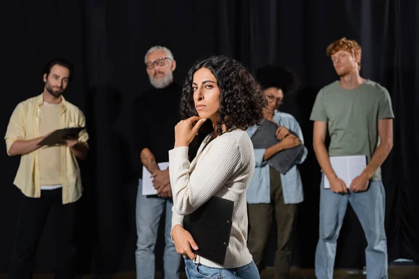 Multiracial woman with strict face expression looking at camera while rehearsing near art director and actors on blurred background - foto de stock