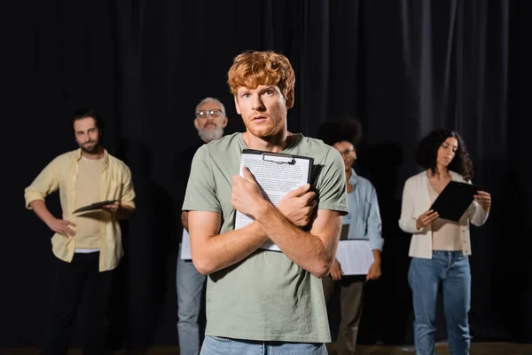 Tensed redhead man holding scenario and looking at camera near multiethnic actors and producer on blurred background - foto de stock