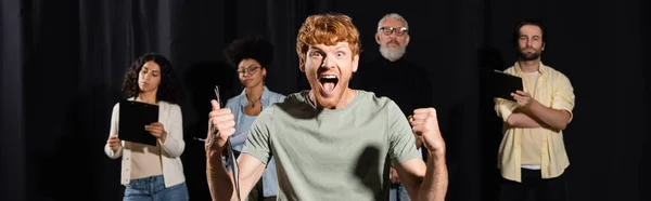 Excited redhead man shouting and showing like gesture near producer and multiethnic actors on blurred background, banner — Fotografia de Stock