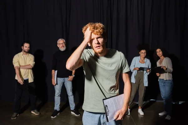 Frustrated redhead man holding clipboard and touching head while looking at camera near actors and producer on blurred background — Foto stock