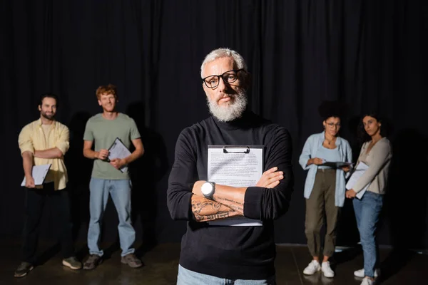Bearded art director standing with crossed arms and screenplay near young multiracial actors. Translation of tattoo: om, shanti, peace — Stock Photo
