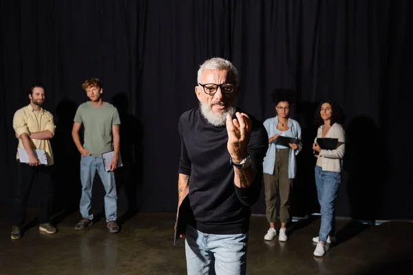 Bearded art director in eyeglasses looking at camera and gesturing near multiracial students on blurred background — Photo de stock