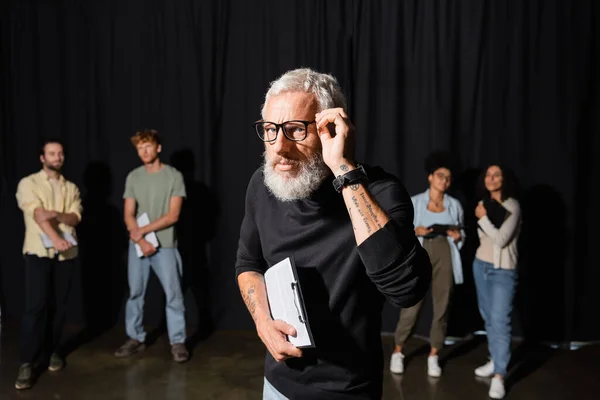 Tattooed teacher adjusting glasses near interracial students. Translation of tattoo: you have to dig deep wells if you want to get clear water — Stock Photo