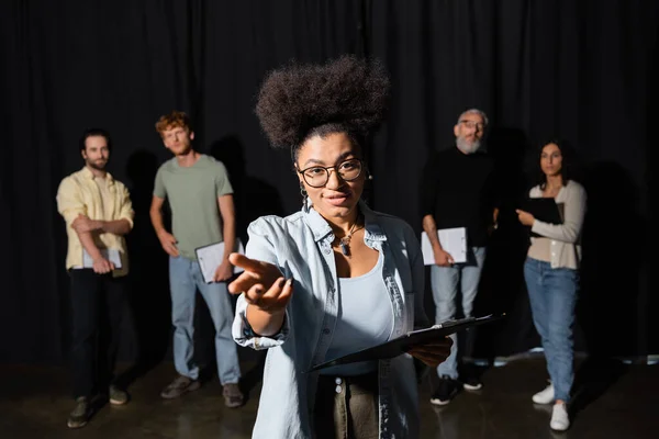 Positive african american woman holding scenario and pointing with hand while rehearsing near blurred interracial students and art director in theater — Foto stock