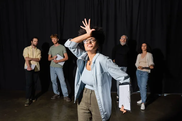 Emotional african american woman grimacing and gesturing near art director and group of multiethnic artists on blurred background — Fotografia de Stock