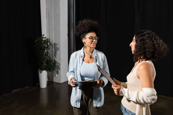 Young multicultural actresses with clipboards smiling at each other in theater — Stock Photo