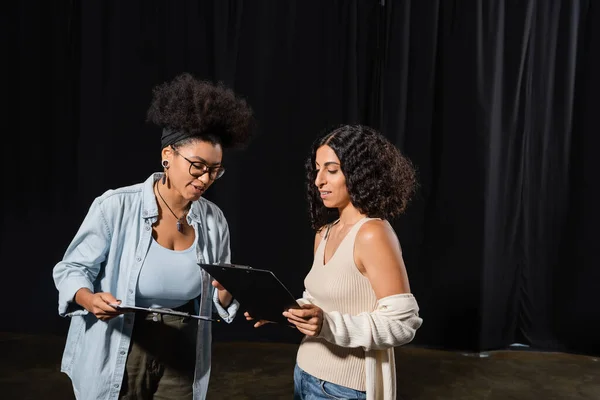 Young interracial actresses looking at clipboards with scenarios in theater — Fotografia de Stock