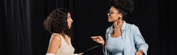 Smiling african american woman in eyeglasses pointing with finger while talking to multiracial actress in theater, banner — стоковое фото
