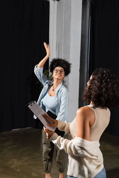 Smiling african american actress in eyeglasses posing with raised hand near multiracial woman with screenplay — Foto stock