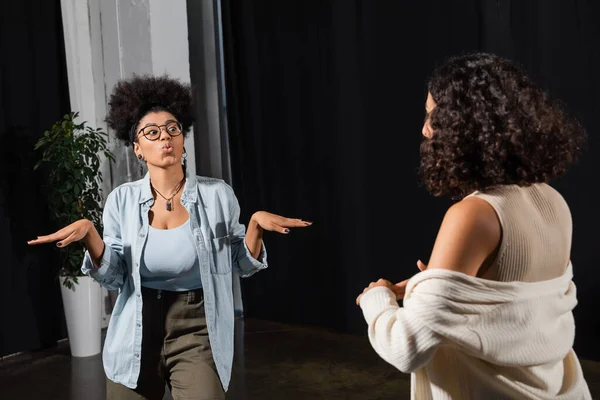 Young african american actress in eyeglasses pouting lips and gesturing while rehearsing near multiracial woman in theater — стокове фото