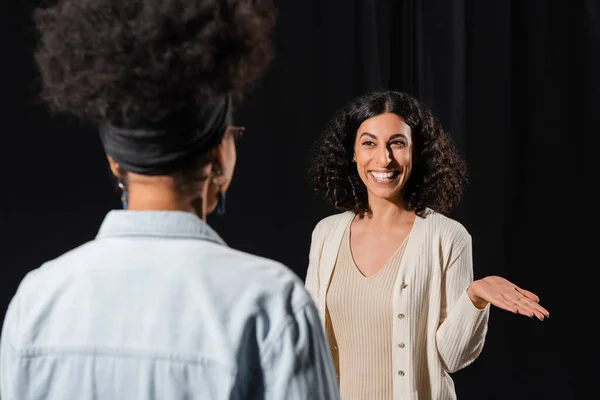 Cheerful multiracial actress pointing with hand while rehearsing near african american woman on blurred foreground — Stock Photo