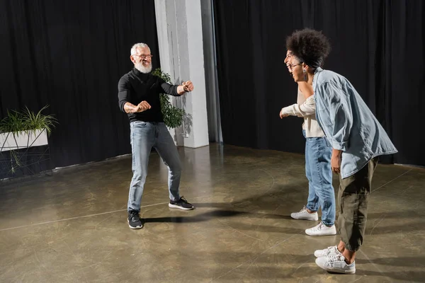 Interracial actresses laughing near bearded art director posing and gesturing in theater. Translation of tattoo: om, shanti, peace — Foto stock